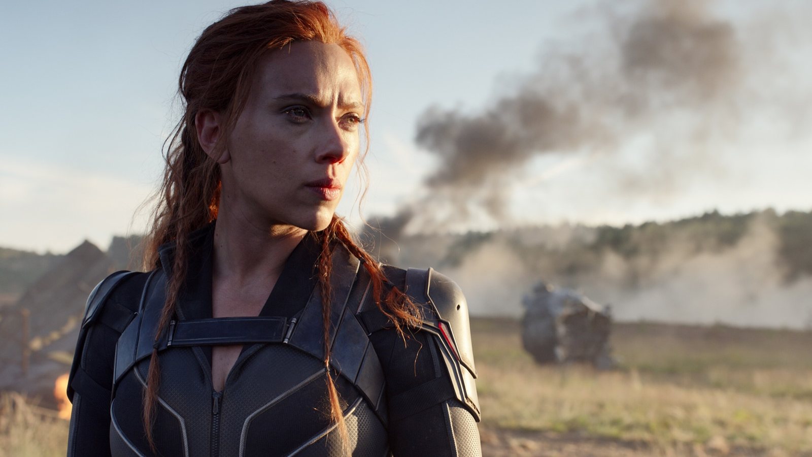 Scarlett Johansson en Natasha Romanoff, en costume, les cheveux au vent devant un champ de bataille enfumé, dans Black Widow